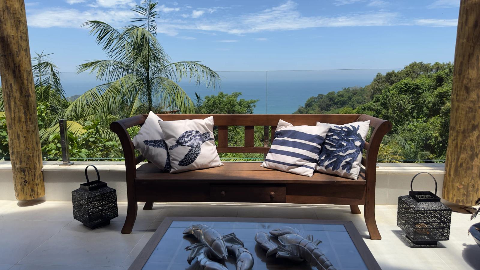 Lounge da varanda da Villa 231 com sofá de madeira e vista para o mar cercada pela Mata Atlântica.